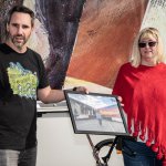 Helmuth Stöber und Künstlerin Brigitte Riedel halten gemeinsam ein Bild. Auf dem Bild sieht man Brigitte vor den 2 LKWs stehen, welche mit ihren Kunstwerken beklebt sind.