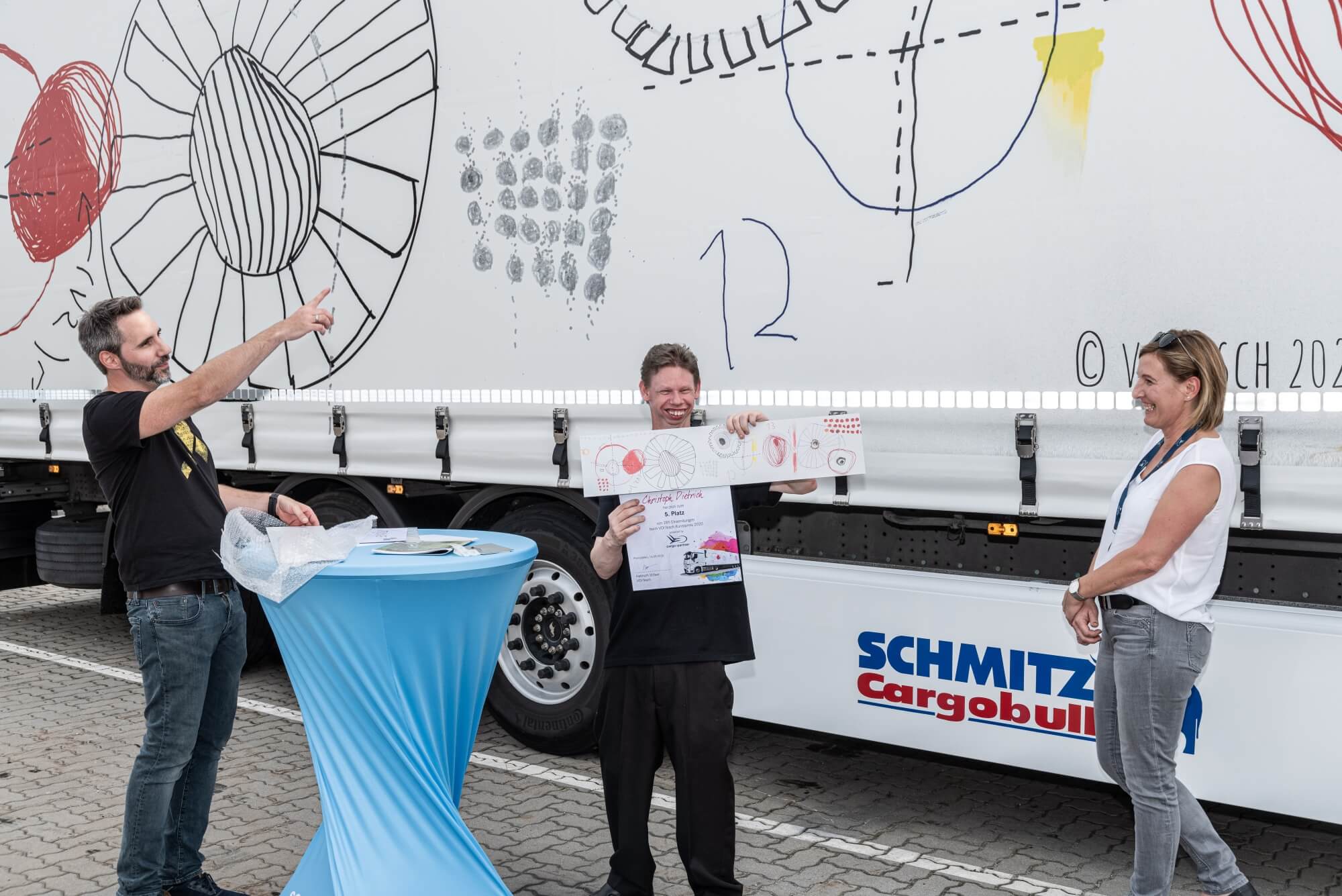 VOI fesch Initiator Helmuth Stöber zeigt auf den LKW mit dem Motiv "Verbindungen". Neben ihm steht der Künstler Christoph Dietrich, welcher das Motiv gemalt hat. Es zeigt kreise und verschiedene geometrische Formen.